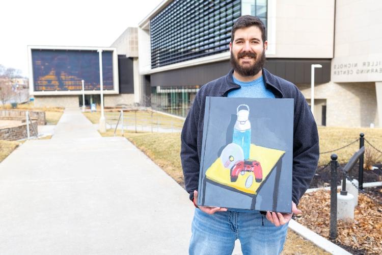 Eric stands outside Miller Nichols Library holding one of his paintings, which depicts a video game controller and military dog tags