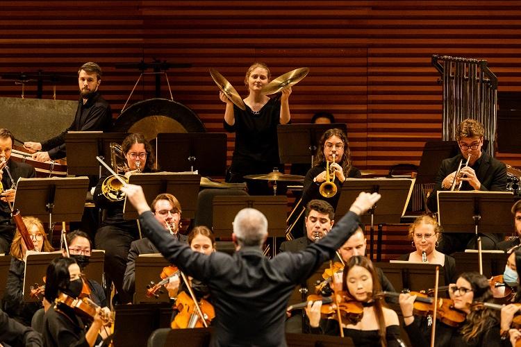 A conductor has his back toward the camera; in front of him is an orchestra performing onstage.