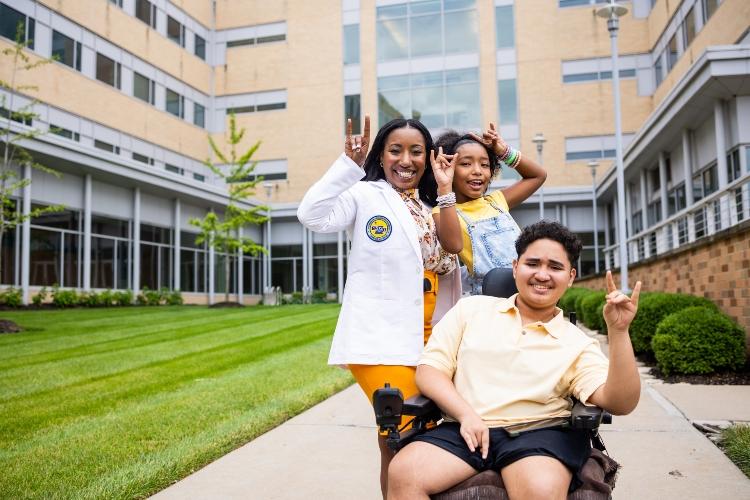 莱蕾萨和她的孩子们笑了, holding up Roo Up hand gestures on the walkway of the Health Sciences building