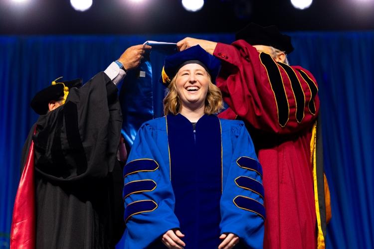 A graduate on stage, smiling as she is hooded for a doctorate degree